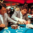 Zwei Männer in grauen Anzügen am Spieltisch, einer hält Karten, der andere ein Getränk, im Casino-Ambiente.