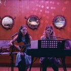 Zwei Frauen musizieren auf einer Bühne, eine spielt Gitarre, die andere Klavier. Im Hintergrund sind Bullaugen an der Wand.