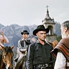 Drei Männer mit Cowboyhüten, zwei auf Pferden, sprechen mit einem älteren Mann. Im Hintergrund sind Berge und ein Kirchturm.