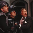 Drei Männer schauen erschrocken in dieselbe Richtung. Einer trägt eine Uniform, der andere einen dunklen Helm.