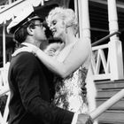 Ich kann keine Personen identifizieren, aber eine Frau in einem glitzernden Kleid umarmt einen Mann in Uniform. Sie sind draußen vor einem Gebäude mit Treppe.