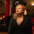 Person mit blondem Haar in Glitzerkleidung vor roten Vorhängen; elegante, stimmungsvolle Beleuchtung.