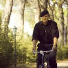 Keine sichtbaren Texte. Ein älterer Mann in Schwarz mit Baskenmütze fährt Fahrrad durch einen lichtdurchfluteten Waldweg.