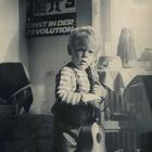 Text: "KUNST IN DER REVOLUTION". Ein Kind mit Streifenpullover hält ein kleines Saiteninstrument in einem Raum.