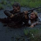 Zwei Personen kämpfen sich durch schlammiges Wasser; eine hält ein Gewehr. Sie sind von Schlamm bedeckt, umgeben von Gras.