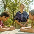 Keine Texte sichtbar. Drei Personen im Freien, zwei Männer sitzen sich gegenüber, eine Frau steht dazwischen, alle sind ernst.