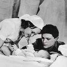 Ein Mann, eine Frau und ein Baby liegen glücklich beisammen auf einem Bett, die Frau trägt eine Haube.