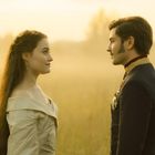 Ein Mann und eine Frau stehen sich in historischer Kleidung gegenüber, vor einem nebligen, goldenen Feldhintergrund.