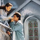 Eine Frau lehnt sich über ein Balkongeländer, ein Mann küsst ihre Hand, romantische Szene bei einem großen Fenster.