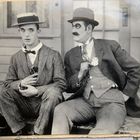 Zwei Männer in Anzügen, ein Mann hält eine Marke, der andere eine kleine Flasche. Beide tragen Hüte.