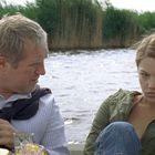 Keine Texte sichtbar. Zwei Personen in einem Boot auf einem Fluss, Schilf im Hintergrund. Eine Person schaut ernst.