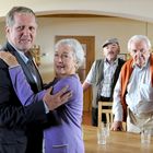 Drei ältere Männer und eine Frau in einem gemütlichen Raum, die Frau tanzt mit einem der Männer.