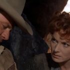 Zwei Personen, ein Mann mit Cowboyhut und eine Frau mit lockigem Haar, blicken ernst drein. Hintergrund ist unscharf.