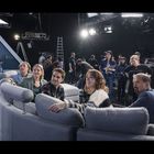 Leute auf grauer Couch, im Studio, Kameras und Equipment, Menschen im Hintergrund, geschäftige Atmosphäre.