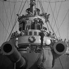 Menschen stehen auf den Decks eines Schiffsturms, viele tragen Uniformen und Kappen, große Kanonenrohre im Vordergrund.