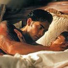 Ich kann den Text im Bild nicht entziffern. Ein tätowierter Mann liegt in einem Bett, hält liebevoll eine Frau und schaut sie an.