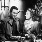 Ein Mann und eine Frau in eleganter Kleidung sitzen an einem Tisch mit Essen und Getränken, sprechen miteinander in stilvollem Ambiente.