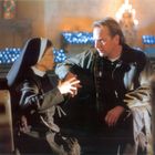 Ein älterer Mann spricht mit einer Nonne in einer Kirche. Im Hintergrund bläuliche Kerzenlichter und ein verschwommenes Kirchenfenster.