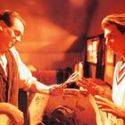 Zwei Männer stehen in einem orange-rot beleuchteten Raum. Einer reicht dem anderen eine Brille, beide scheinen intensiv zu diskutieren.