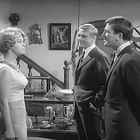 Ein Raum mit Gemälden und Dekorationen; drei Personen, zwei Männer im Anzug sprechen mit einer Frau.