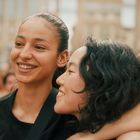 Zwei lachende Menschen, eng umarmt, in einer fröhlichen Menge. Hintergrund unscharf, Gebäude erkennbar.