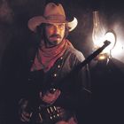 Ein Mann im Cowboyhut steht mit Gewehr in einem dunklen Raum neben einer Öl-Lampe. Er hat einen roten Schal um den Hals.