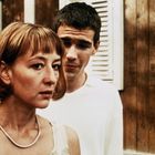 Zwei Personen stehen vor einem Holzhaus mit weißen Fensterläden. Eine Frau im Vordergrund schaut ernst nach links.