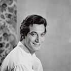 Ein Mann mit dunklem Haar lächelt, trägt ein helles Hemd, mit dekorativem Hintergrund auf der linken Seite. Keine erkennbaren Texte.