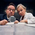 Zwei Personen in formeller Kleidung betrachten etwas mit einer Lupe auf einem Tisch. Rechts liegt ein geöffnetes Buch.