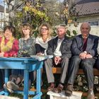 Eine Gruppe von sieben Menschen sitzt im Garten auf einer Bank neben einem blauen Tisch mit Äpfeln und Trauben.