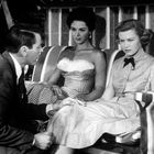 Zwei Frauen sitzen auf einer Schaukel, eine in einem karierten Kleid, die andere in einem Hemd mit Fliege, neben einem Mann.