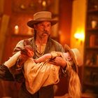 Ein Mann in einem braunen Hut trägt ein junges Mädchen in einem beigefarbenen Kleid und Strohhut in einem warm beleuchteten Raum.