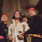 Zwei Männer mit roten Stirnbändern halten eine Frau in einem weißen Hemd fest. Sie hat ein entschlossenes Gesicht.