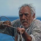 Ein älterer Mann mit grauen Haaren zieht mit angestrengtem Gesichtsausdruck ein Seil vor einem verschwommenen Meereshintergrund.