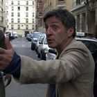 Drei Personen in einer engen Stadtstraße; ein Mann gestikuliert intensiv, eine Frau schaut überrascht, ein anderer Mann sieht zu.