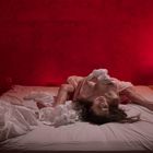 Ein spärlich beleuchteter Raum mit rotem Licht. Eine Person auf einem Bett mit weißen Laken, Lampe rechts.