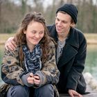 Ein junger Mann lächelt und legt den Arm um eine junge Frau, die schüchtern lächelt. Sie sitzen im Freien an einem Gewässer.