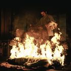 Ich kann keine Person erkennen. Ein Mann steht in einem dunklen Raum und starrt auf ein loderndes Feuer auf dem Boden.