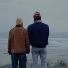 Zwei Menschen stehen am Strand, schauen auf das Meer. Einer trägt eine gelbe Kapuze, der andere eine rote Mütze.