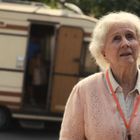 Eine ältere Frau schaut nachdenklich nach oben. Im Hintergrund steht ein Wohnwagen mit geöffneter Tür.