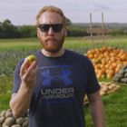 Der Mann trägt ein "Under Armour"-Shirt, Sonnenbrillen und hält einen Apfel in einer landwirtschaftlichen Umgebung.
