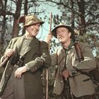 Zwei Männer in traditioneller Tracht, lächeln in bewaldeter Umgebung. Einer hält ein Fernglas. Beide tragen Hüte.