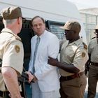 Ein Mann im weißen Anzug wird von drei Wachmännern in beige Uniformen zu einem Gefängnisfahrzeug begleitet.