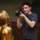 Eine Person mit Kamera filmt eine mit goldener Farbe bemalte Person in einem Raum mit Holzverkleidung.
