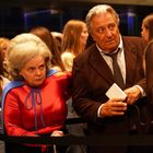 Ein älterer Mann im Anzug und eine Frau im Superheldenkostüm stehen in einer Menschenmenge und schauen ernst.