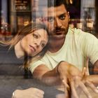 Ein Paar sitzt am Tisch; die Frau lehnt ihren Kopf auf die Schulter des Mannes. Eine Tasse steht vor ihnen.