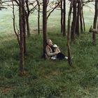 Ein älterer Mann und ein Kind sitzen umgeben von Bäumen im Gras, der Mann umarmt das Kind liebevoll.