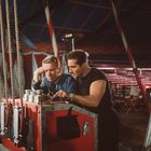 Zwei Männer stehen an einem Konstrukt aus Metall und Seilen in einem Zirkuszelt. Sie wirken konzentriert und beraten sich.