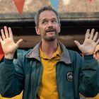 Auf einer Jacke steht "Camp Corp". Ein Mann hebt lächelnd die Hände, umgeben von Menschen, bunte Wimpel im Hintergrund.