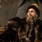 Ein Mann in opulenter Kleidung mit Pelz, Krone und Zepter sitzt vor einem steinernen Hintergrund.